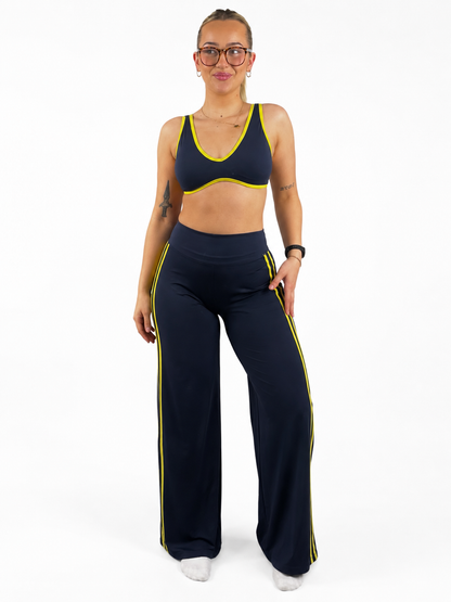 A woman modeling a matching navy blue activewear set with bright yellow trim, consisting of a sports bra and wide-leg flares.