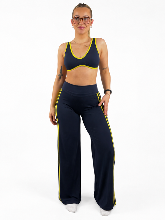A woman modeling a matching navy blue activewear set with bright yellow trim, consisting of a sports bra and wide-leg flares.